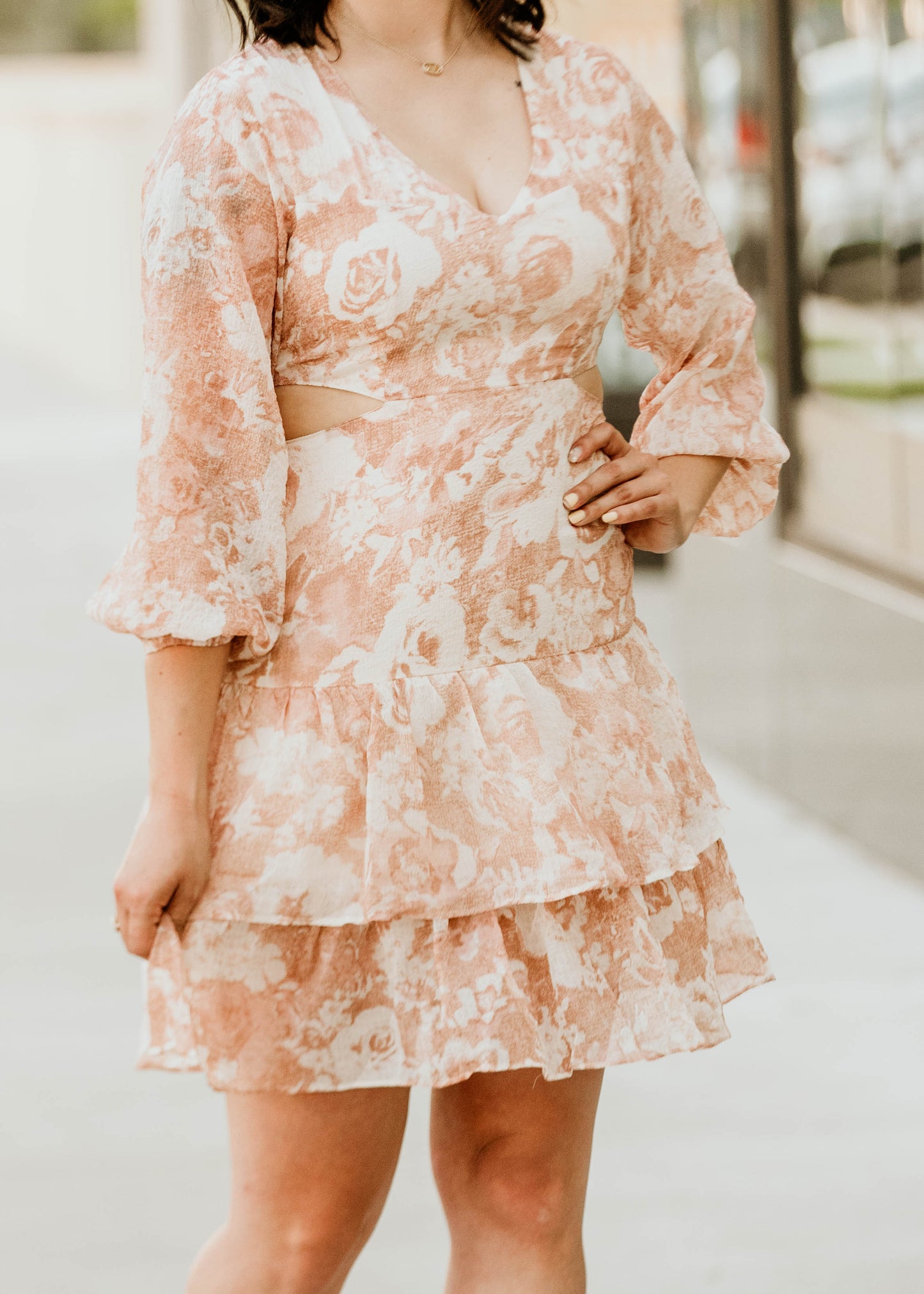 A flowy white dress adorned with soft, light burnt orange floral patterns, creating a fresh and vibrant look. It features stylish cut-outs on the sides for a modern touch, and a flattering V-neckline at the chest for added elegance. The dress falls right above the knee, offering a perfect balance of comfort and chic, ideal for casual outings or a daytime event.