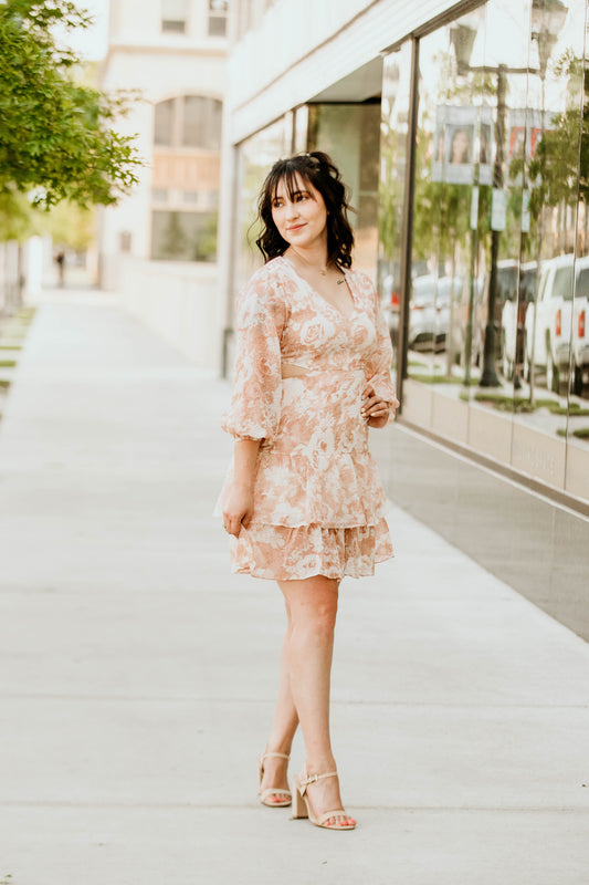 A flowy white dress adorned with soft, light burnt orange floral patterns, creating a fresh and vibrant look. It features stylish cut-outs on the sides for a modern touch, and a flattering V-neckline at the chest for added elegance. The dress falls right above the knee, offering a perfect balance of comfort and chic, ideal for casual outings or a daytime event.